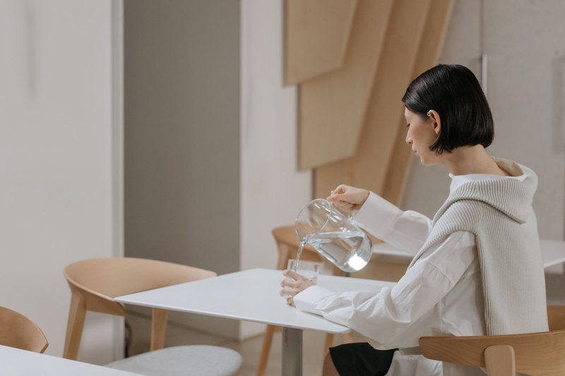 A glass of water being poured from a pitcher onto a light-colored table.