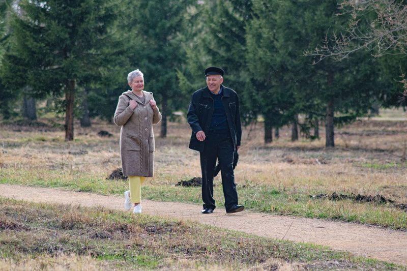 Two older adults walking together along a tree-lined park path in daylight
